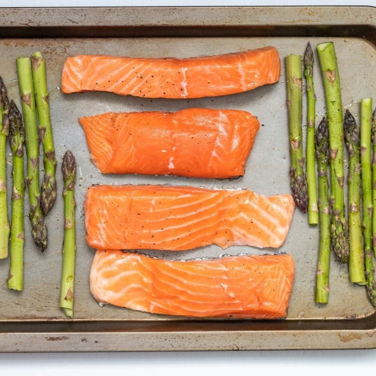 Four salmon fillets lined up on a baking tray, book-ended by asparagus spears