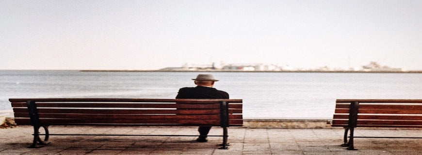 elderly man sitting alone on bench, his back to the camera lens, staring across the water