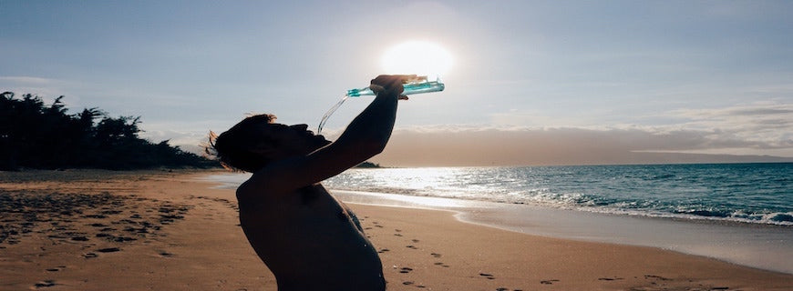 shirtless man on sun-kissed beach, tipping a water bottle into his mouth