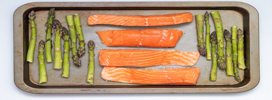 Four salmon fillets lined up on a baking tray, book-ended by asparagus spears