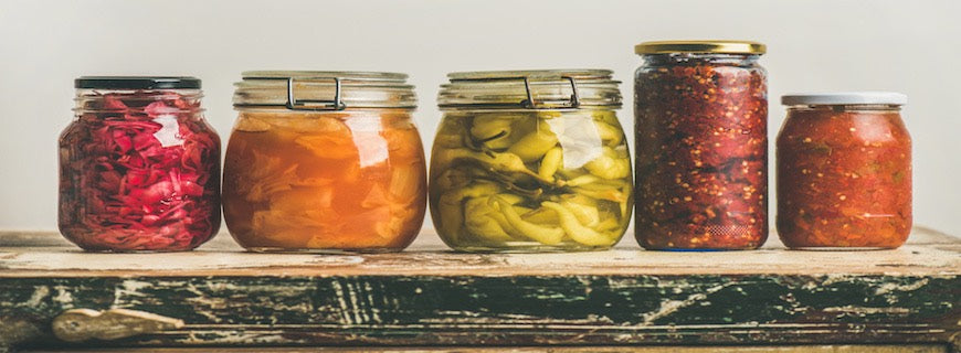 Pickled/fermented vegetables in jars placed in row over vintage kitchen drawer, against white wall background.